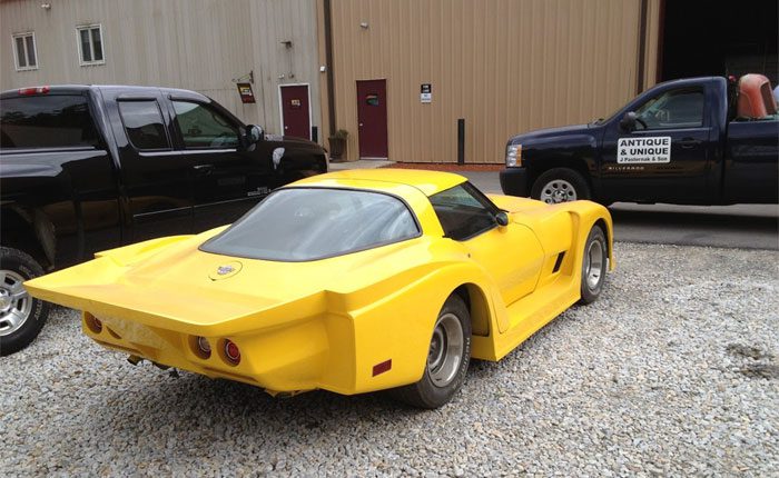 A bright yellow vintage sports car with a custom body parked next to antique-themed trucks on a gravel road.