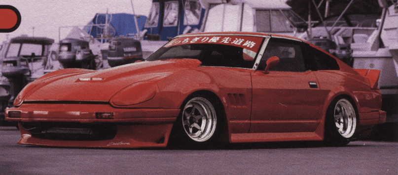 A retro-styled red sports car with Japanese text on the windshield parked in front of a marina with boats in the background.