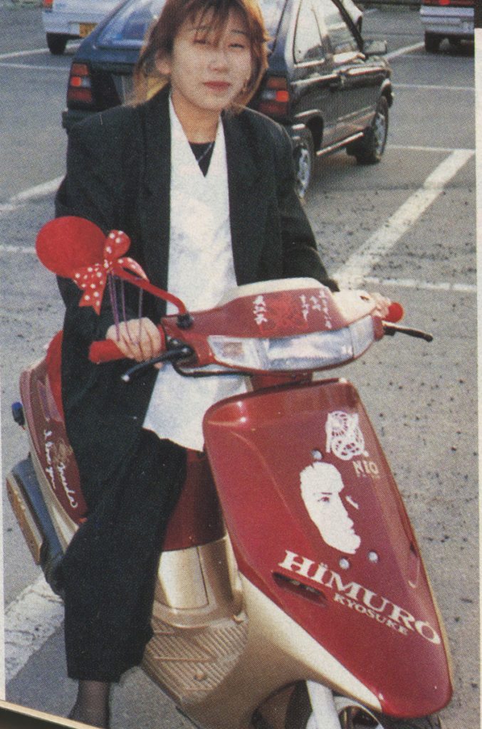 Young woman riding a red Honda Dio AF18 scooter with HIMURO and KYOUSUKE Japanese text, and a decorative ribbon in a parking lot.