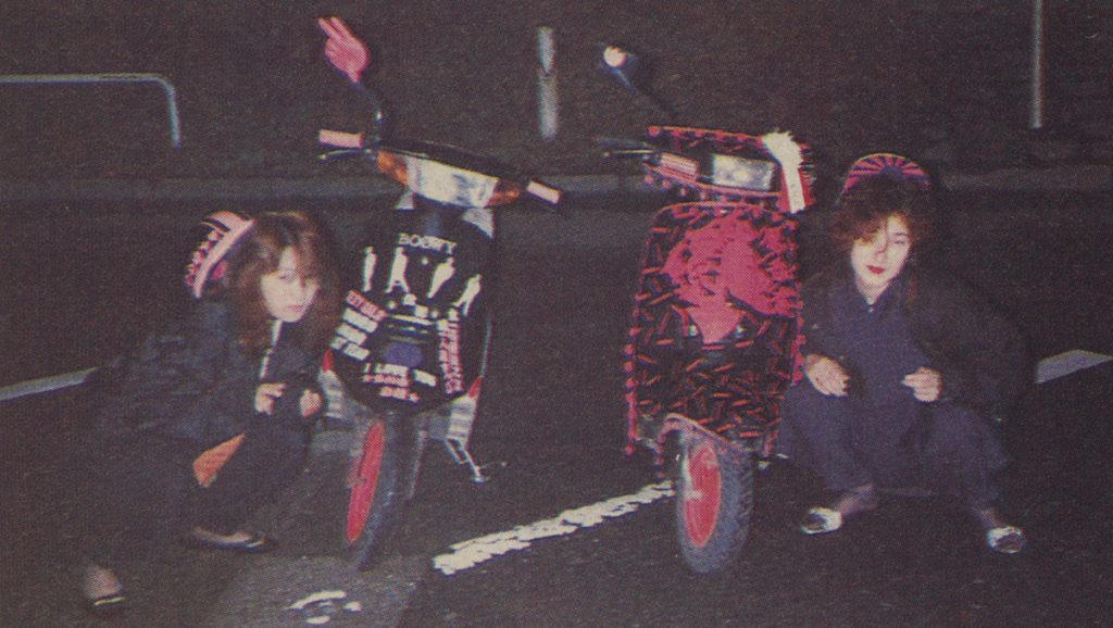 Two women squatting next to vibrantly decorated scooters at night. One scooter has 'ROCKY' on it. 1980s Japan.