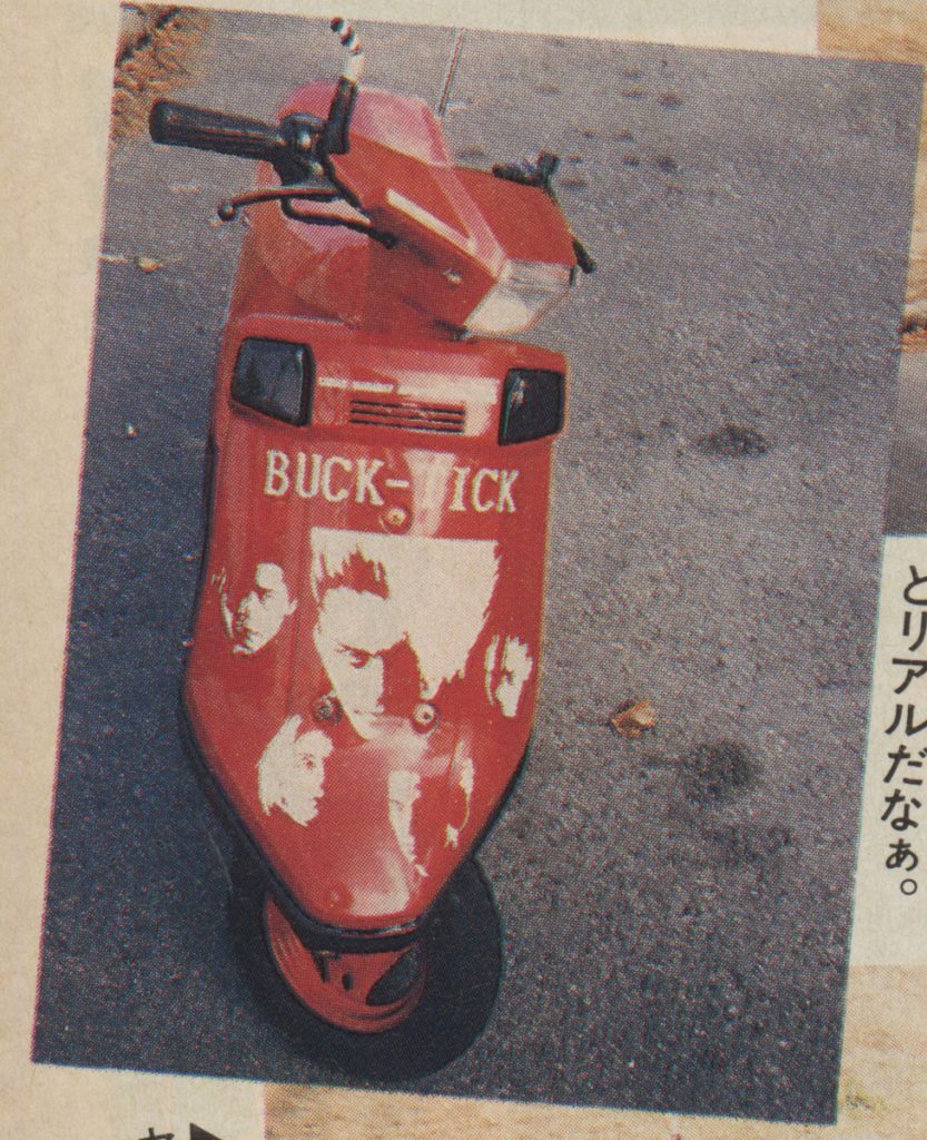 Red scooter with BUCK-TICK and band members' images on the front, parked on pavement. Japanese text: とリアルだなぁ on the right.