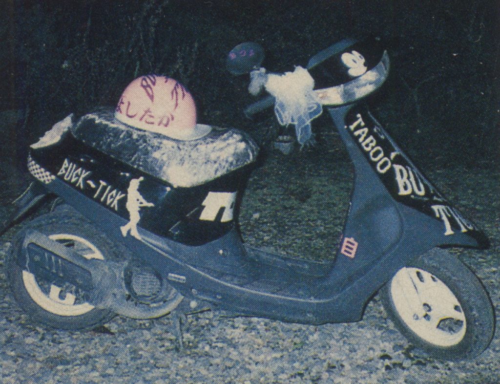 Decorated scooter with BUCK-TICK and TABOO text, and a pink helmet with Japanese writing, in a night setting.