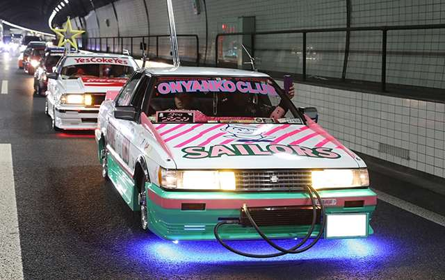 Colorful custom cars with a Toyota Mark II GX71 with neon underglow lights and decorations driving through a tunnel in a lively car parade.