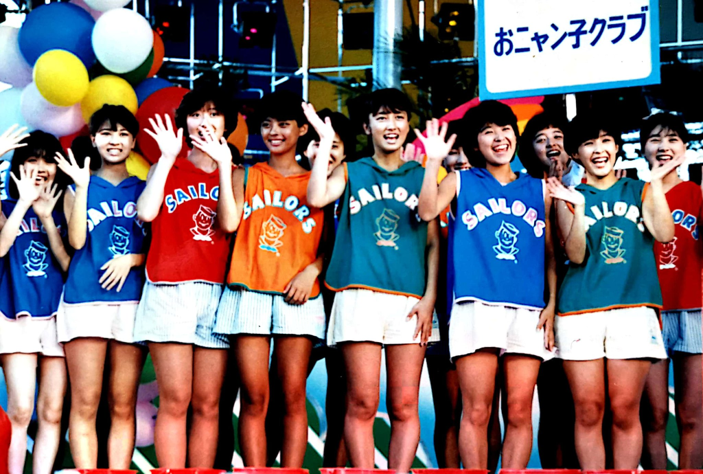 Group of smiling girls in colorful Sailors uniforms waving, with balloons in the background at a festive event.