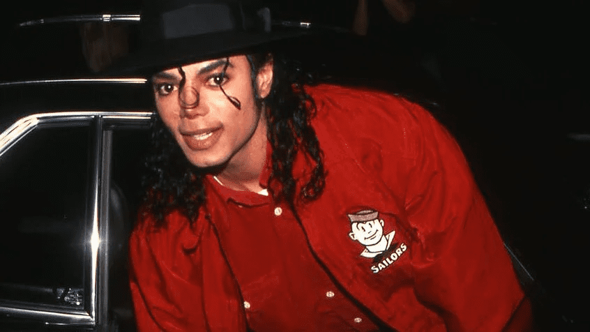 Michael Jackson wearing a red Sailors jacket and black hat with curly hair, smiling, and standing next to a car.