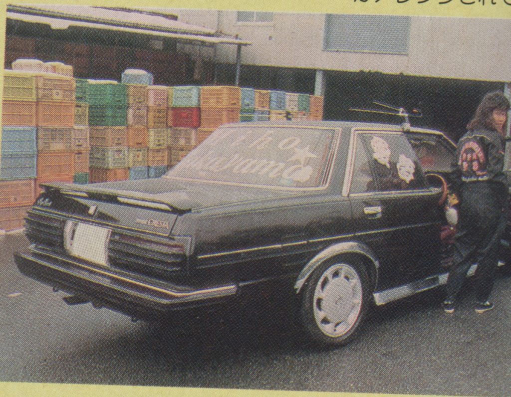 Retro black car with custom decals parked near colorful crates, person in black jumpsuit standing beside.