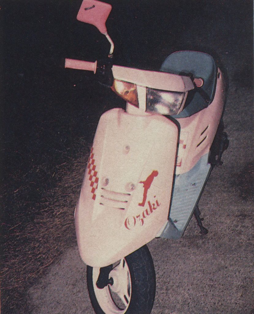 Vintage Yamaha Jog scooter with a pink and white Ozaki color scheme and checkered detailing parked on a dark street.