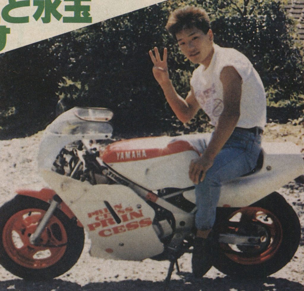 Young man sitting on a Yamaha motorbike with custom graphics, making a peace sign with nature in the background.