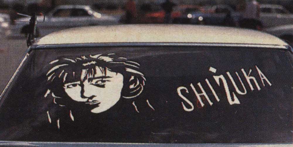 Car with a rear window decal of a woman's face and the word Shizuka in bold letters, parked in a busy lot.