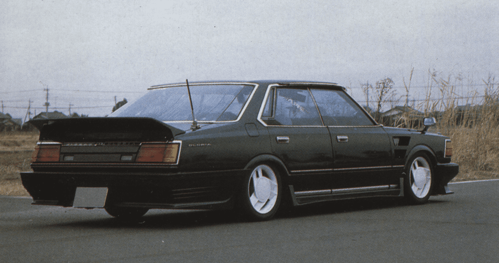 A black vintage Nissan Gloria 430sedan with a spoiler, low stance, and white wheels on a deserted road.