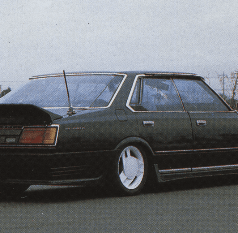 A black vintage Nissan Gloria sedan with a spoiler, low stance, and white wheels on a deserted road.