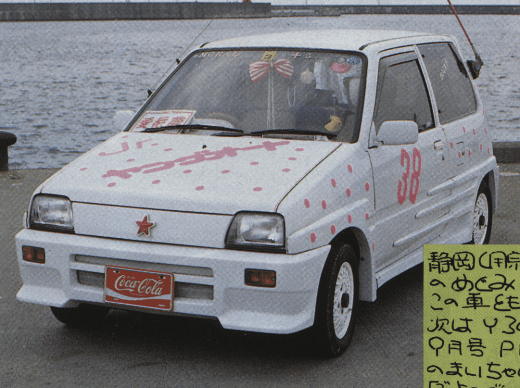 White car with pink polka dots and a Coca-Cola license plate parked by the sea. 