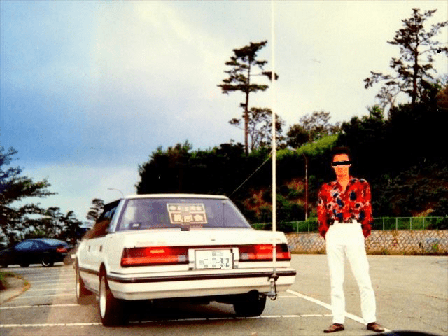 A man in colorful shirt stands beside a vintage Japanese car in a scenic parking lot surrounded by trees.