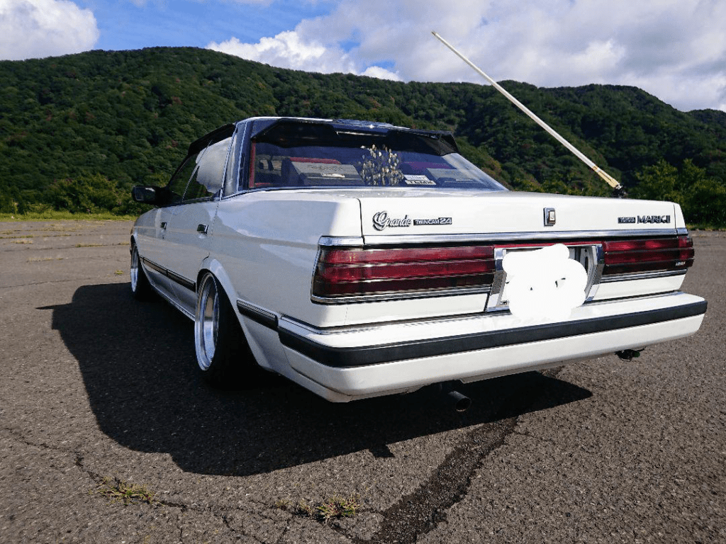 Rear view of a white Toyota Mark II Grande, parked on pavement with a scenic, green mountain backdrop on a sunny day.