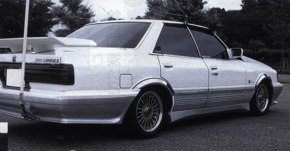 Rear view of a modified Toyota Mark II with a custom spoiler and rims, parked outdoors in a scenic area.