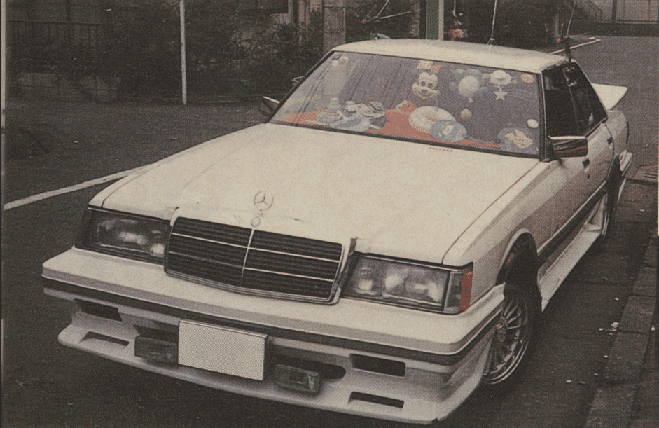 White customized Toyota GX61 Mark ii with Japanese license plate, Mercedes-Benz 190e grille, and toys on the dashboard parked on a city street.