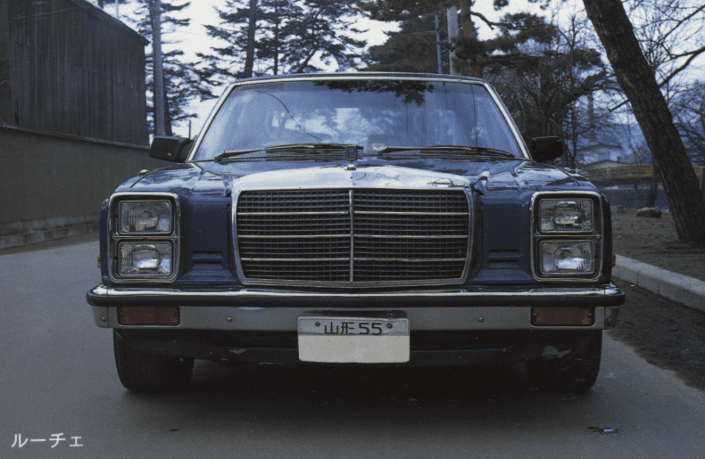 Front view of a blue vintage Japanese car Mazda Luce LA4 with the brand name Luce written in Japanese katakana on the bottom left.