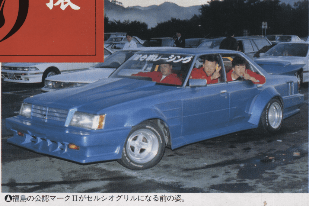 Blue modified Toyota GX61 Mark ii car with three people in red jackets at an event in Fukushima, Japan; windshield text reads ドリフトレーシング (Drift Racing).