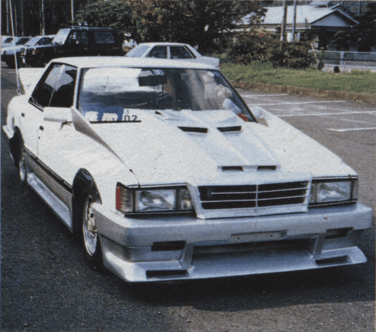 Modified white classic Toyota Mark ii car with opaque license plate parked in front of trees on a sunny day. Japanese text translates to Carnet.