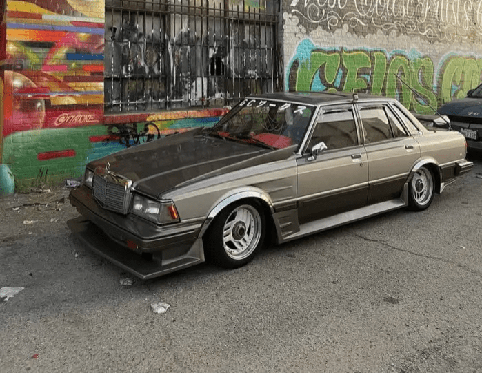 Vintage modified car parked in front of colorful graffiti wall, showcasing custom rims and lowered stance.