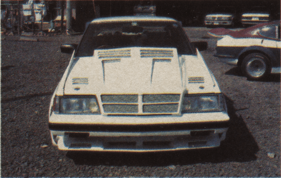 Front view of a modified white Toyota GX61 Mark ii sports car with hood vents in a gravel parking lot, with cars in the background.