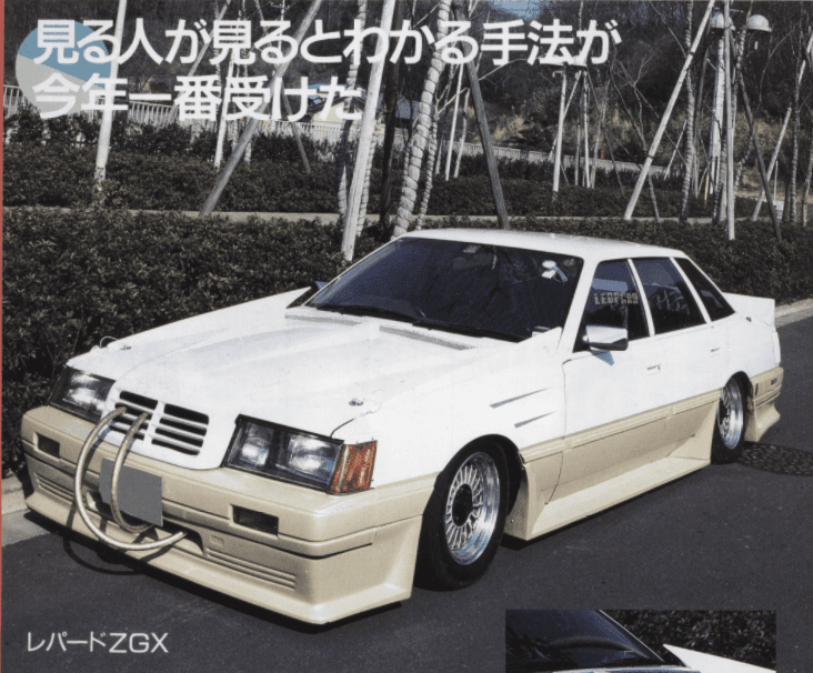 A Haiso customized white Nissan Leopard ZGX car with modified bumper and low stance, parked on a street with trees in the background. White fiberglass mercedes style grille with aftermarket hood and fender spoilers.