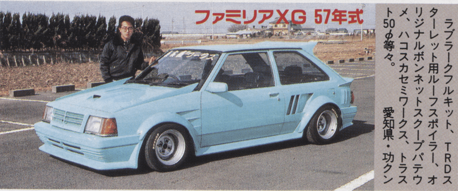 Vintage blue Mazda Familia XG with custom body kit parked on an asphalt lot with a person standing beside it in Japan.