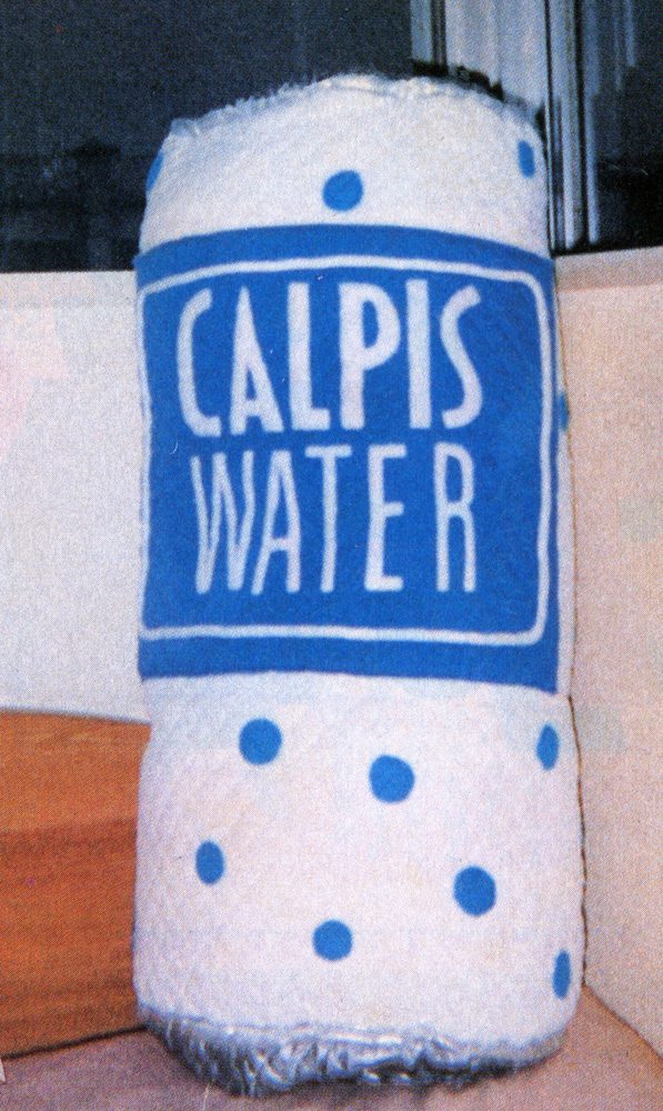 Oversized plush toy of a Calpis Water bottle with blue polka dots, displayed indoors.