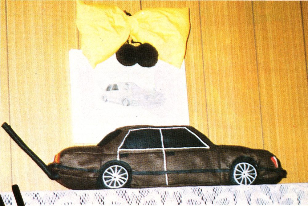 Soft toy car on shelf with wall art of a car and large yellow bow hanging above, creating a whimsical display.