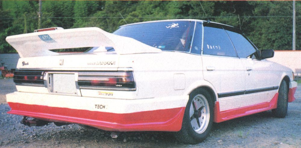 Vintage white car with red trim and large rear spoiler, parked on gravel, featuring Japanese decals on rear window.