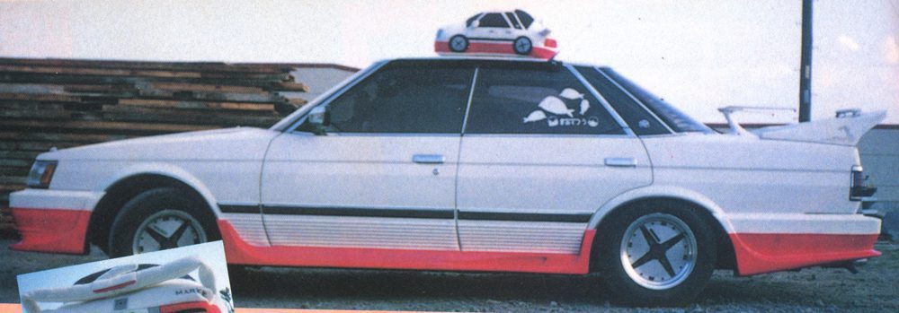 Retro modified car with toy car on roof, featuring unique decals and red accents, parked outdoors.