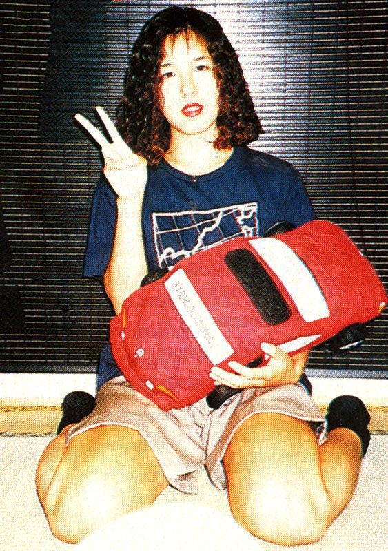 Person holding red car pillow, making peace sign, wearing blue shirt with map design. Cozy indoor setting.