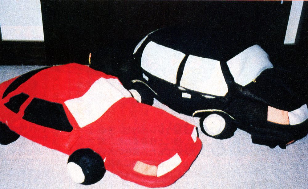 Two plush toy cars in red and black colors on a carpet floor, designed with detailed windows and wheels.