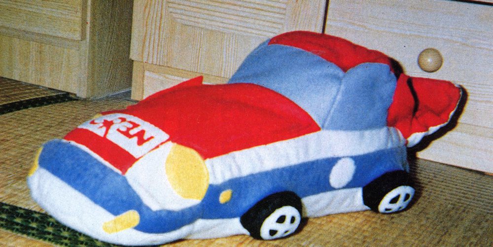 Colorful car-shaped plush toy with Japanese text ネコ (neko) meaning cat on a tatami floor near wooden furniture.