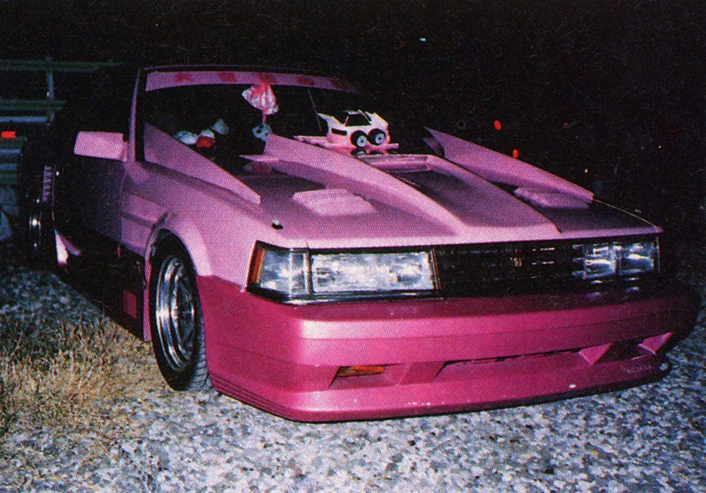 Pink modified car at night with cartoon sticker on windshield in a rocky area. Japanese text reads Kaido meaning highway.