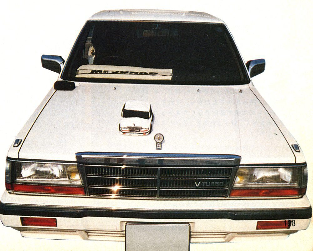 1980s white V turbo sedan with mini car model on hood, windshield displaying M.Yurikago text.