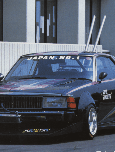 Modified vintage Japanese race car with custom decals and tall exhaust pipes, parked in front of a modern building.
