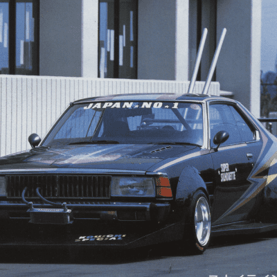 Modified vintage Japanese race car with custom decals and tall exhaust pipes, parked in front of a modern building.