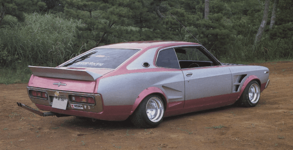 Vintage custom car in a forest setting, featuring a bold pink and silver paint job, wide tires, and a rear spoiler.