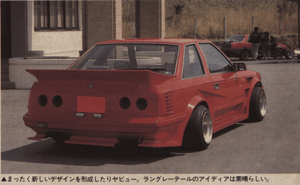 Red modified sports car with wide rear wing parked near building.