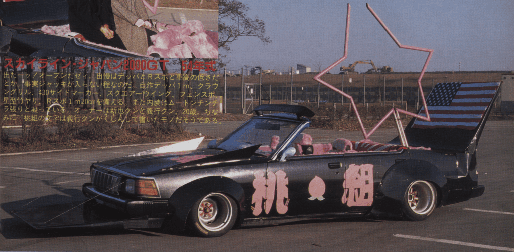 Custom car with pink interior, star-shaped frame, and American flag design, parked in an industrial area.