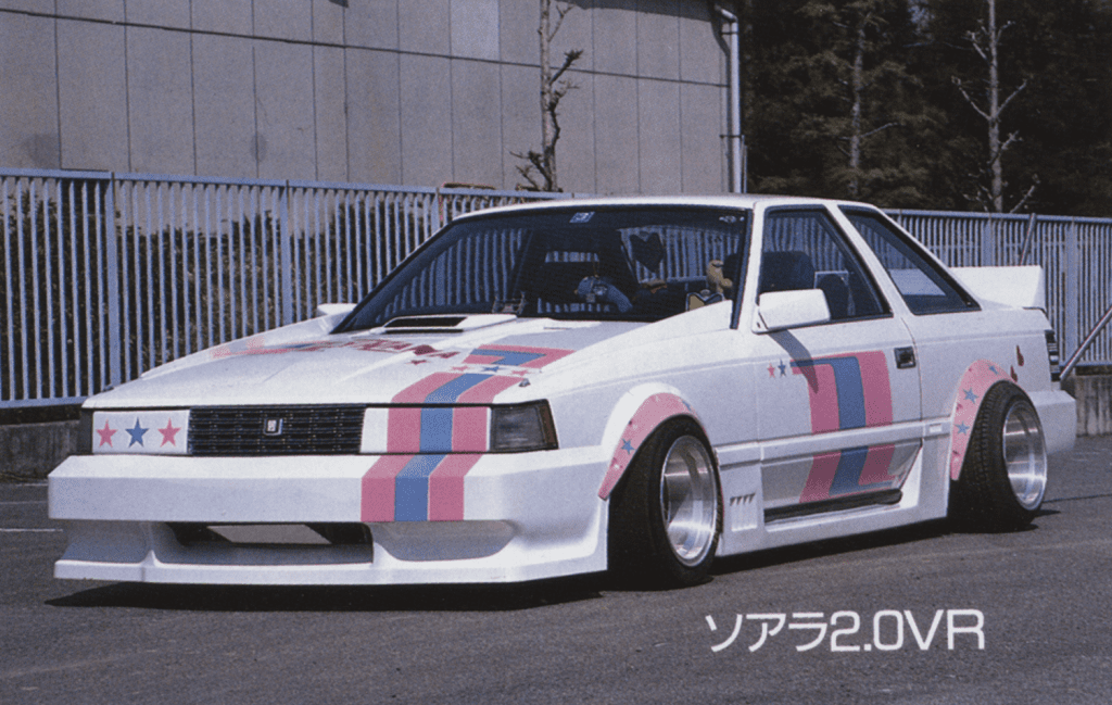 Vintage modified car with white body and pink-blue racing stripes parked near a fence; retro design elements visible.