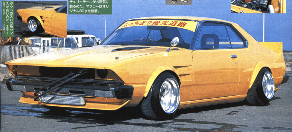 Vintage yellow car with custom modifications and a spoiler, parked outdoors under a clear blue sky.