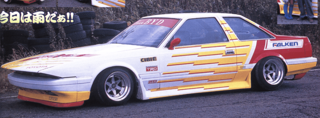 Custom white and yellow retro sports car with decals parked on a road with tires in the background.