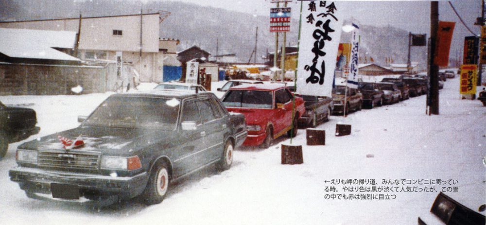 Vintage cars lined up in snowy street, Japan, with traditional banners, creating a nostalgic winter scene.