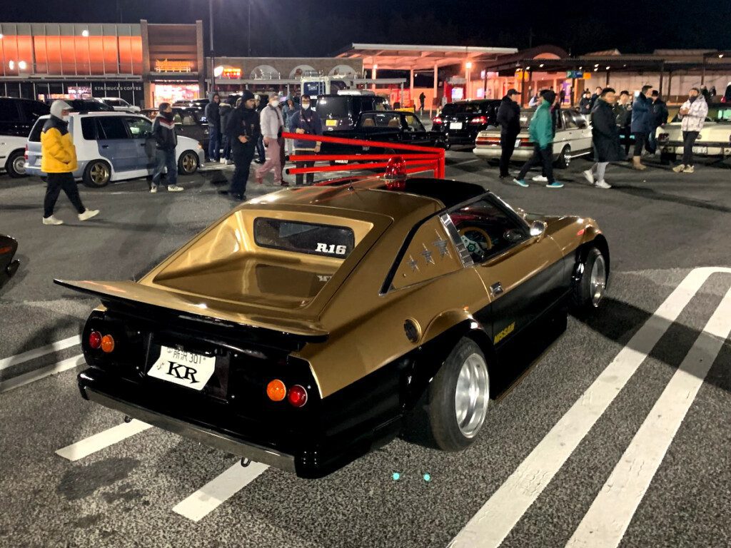 Custom retro car at a night car meet in a busy parking lot with people gathered around.