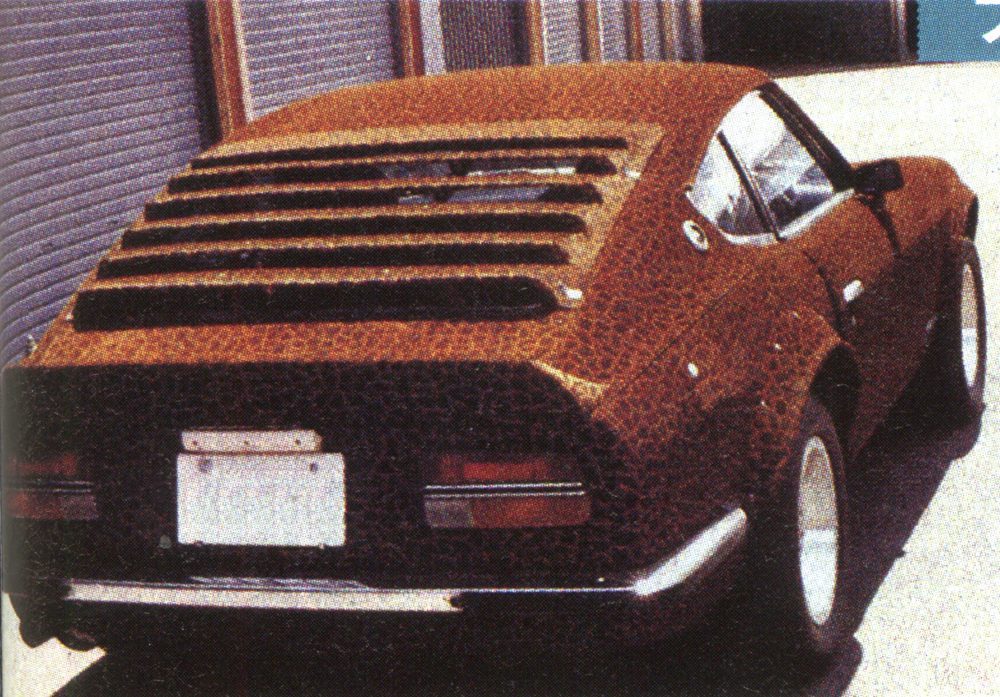 Vintage sports car with louvers, custom leopard print paint, parked near roll-up doors on a sunny day.