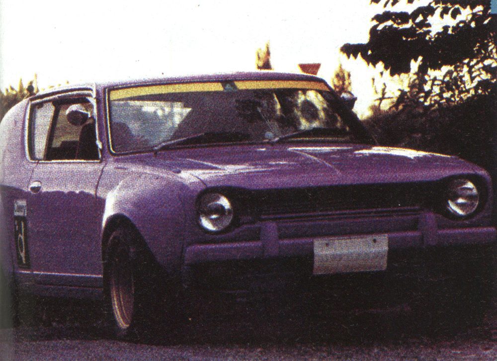 Vintage purple car parked on a street, surrounded by trees, in a sunset setting.