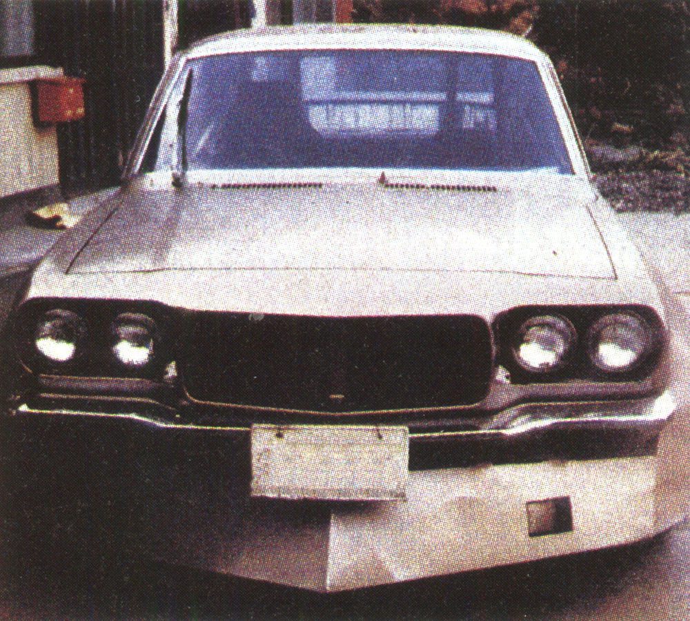 Vintage car with custom front plate and chrome bumper in a driveway, featuring dual headlights and unique grill design.