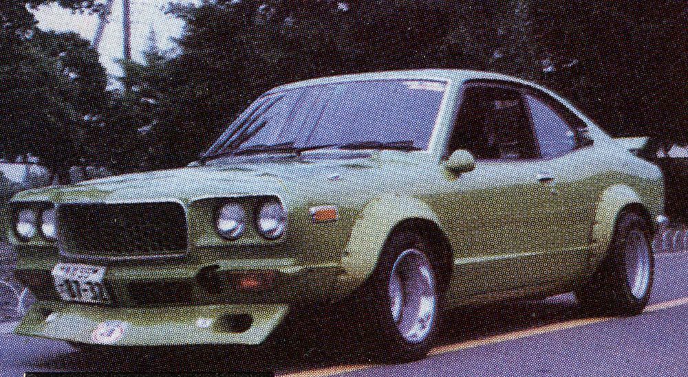 Vintage green sports car with custom modifications on a road, surrounded by trees.